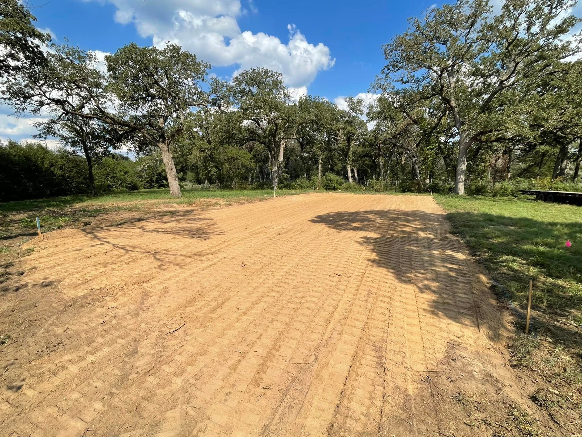 Cleared dirt area in a field, surrounded by trees, under a blue sky with clouds.