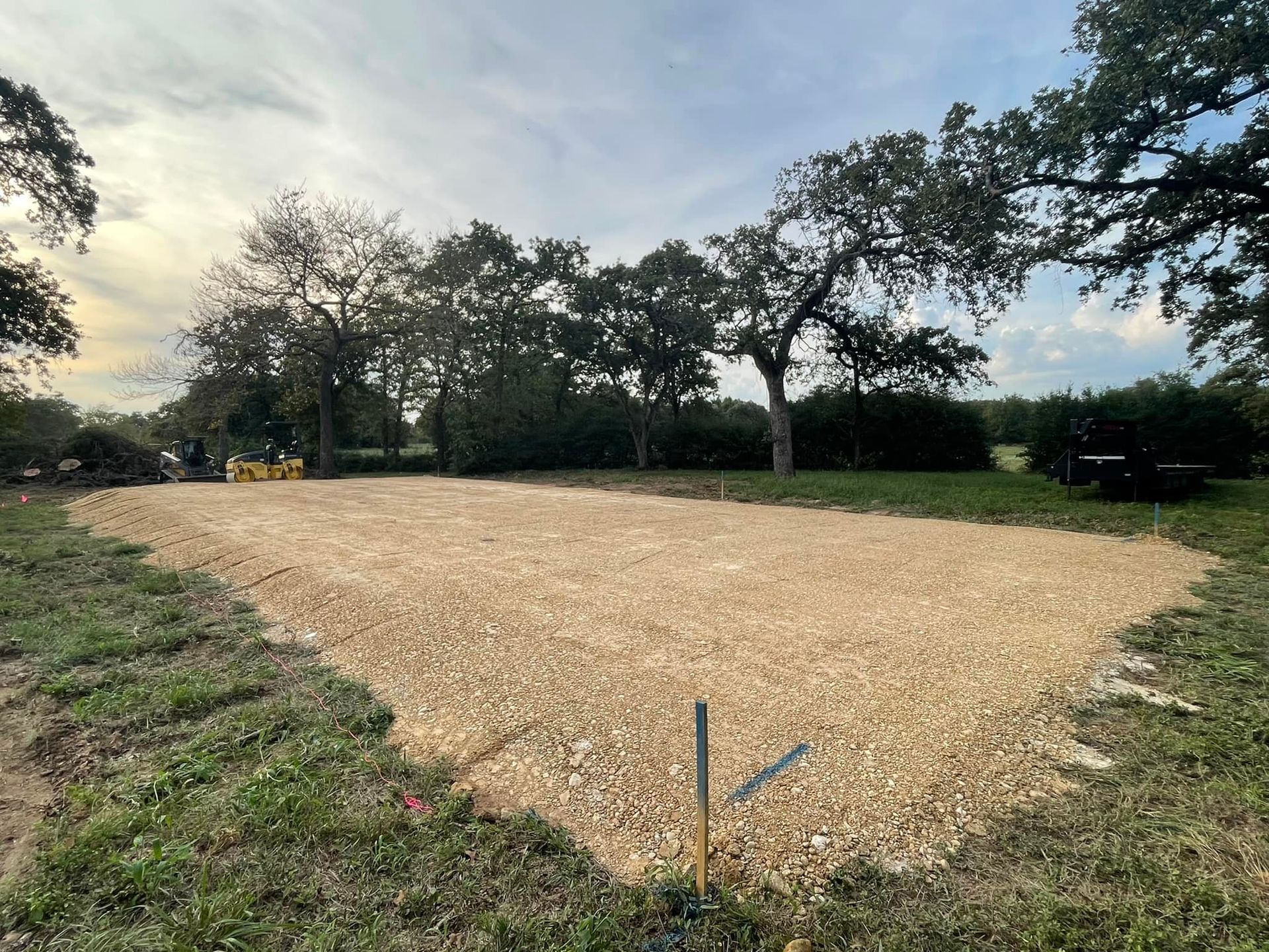 A gravel pad prepared for construction, surrounded by grass and trees under a cloudy sky.