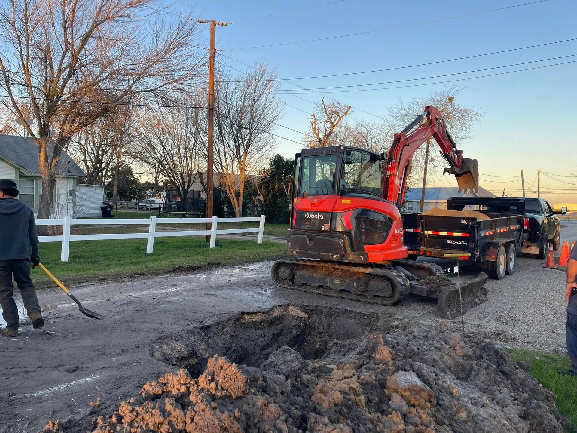 An excavator and workers on a road excavation project. The red excavator is loading dirt into a trailer.