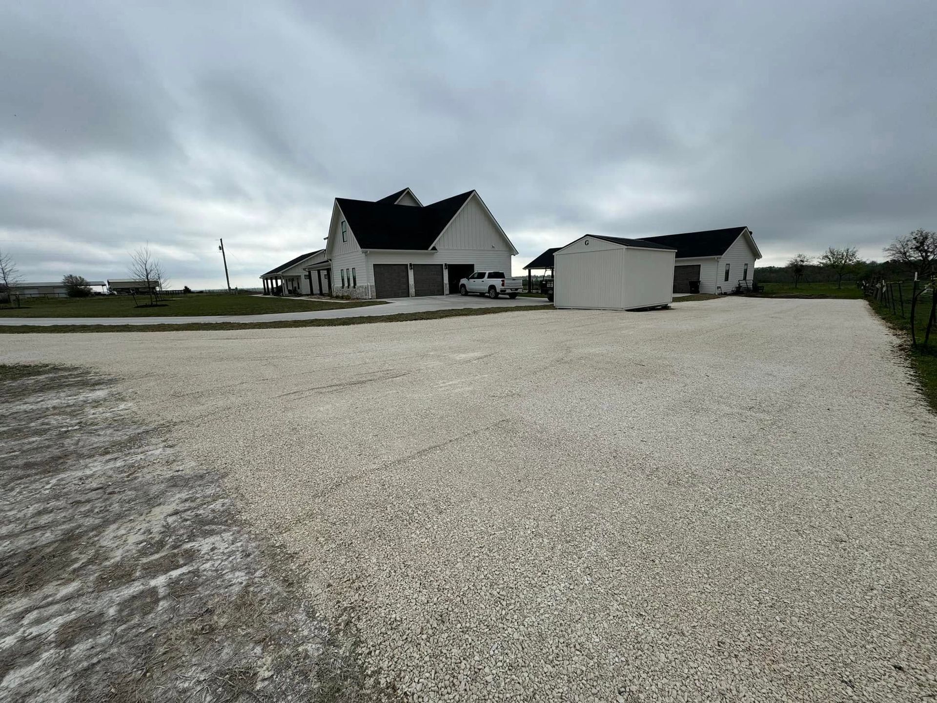 Gravel driveway leading to a white house with a black roof, under a cloudy sky.