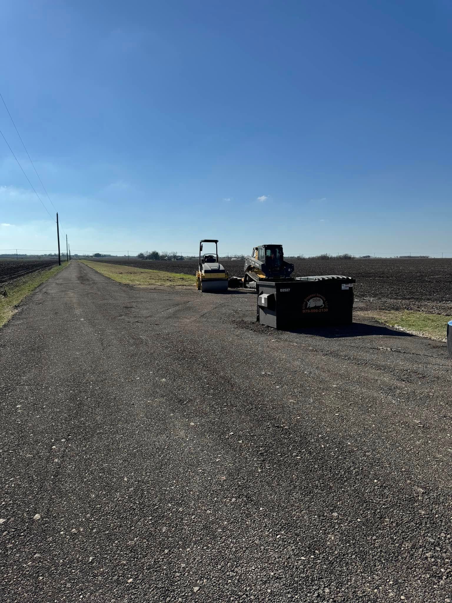 Gravel road through farmland; small vehicles and equipment sit along the roadside under a blue sky.