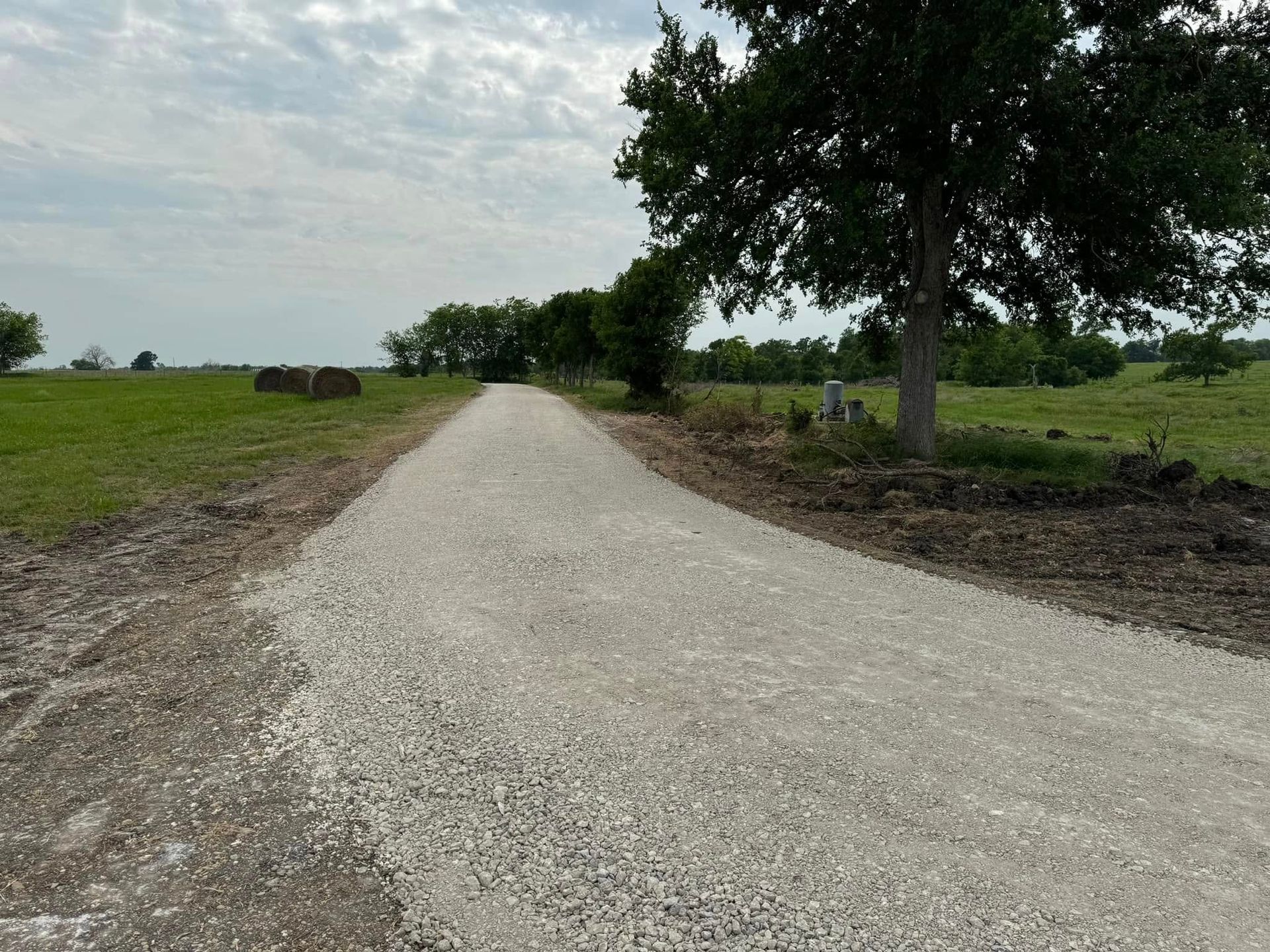 Gravel road through a green field, trees on the right, bales of hay on the left, overcast sky.