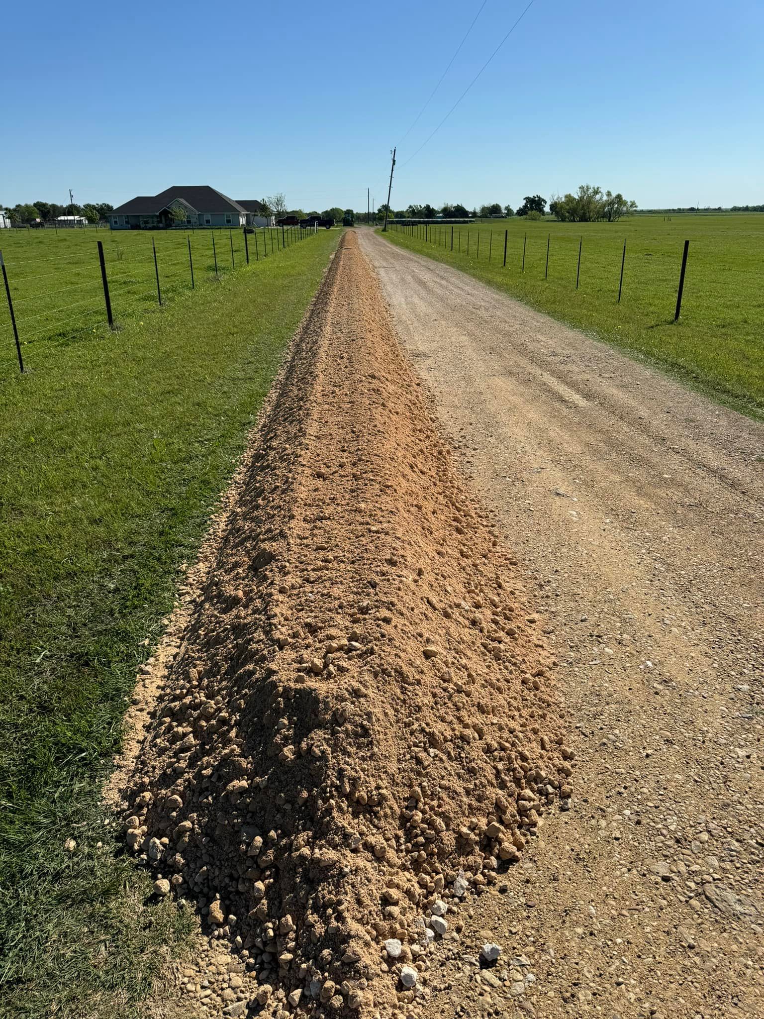 Gravel road through a grassy field on a sunny day, leading to a house in the distance.