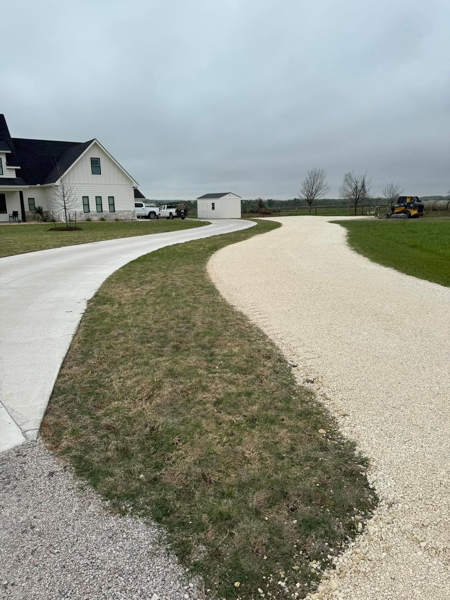 A long, winding driveway with gravel and concrete paths leads to a white farmhouse on a cloudy day.