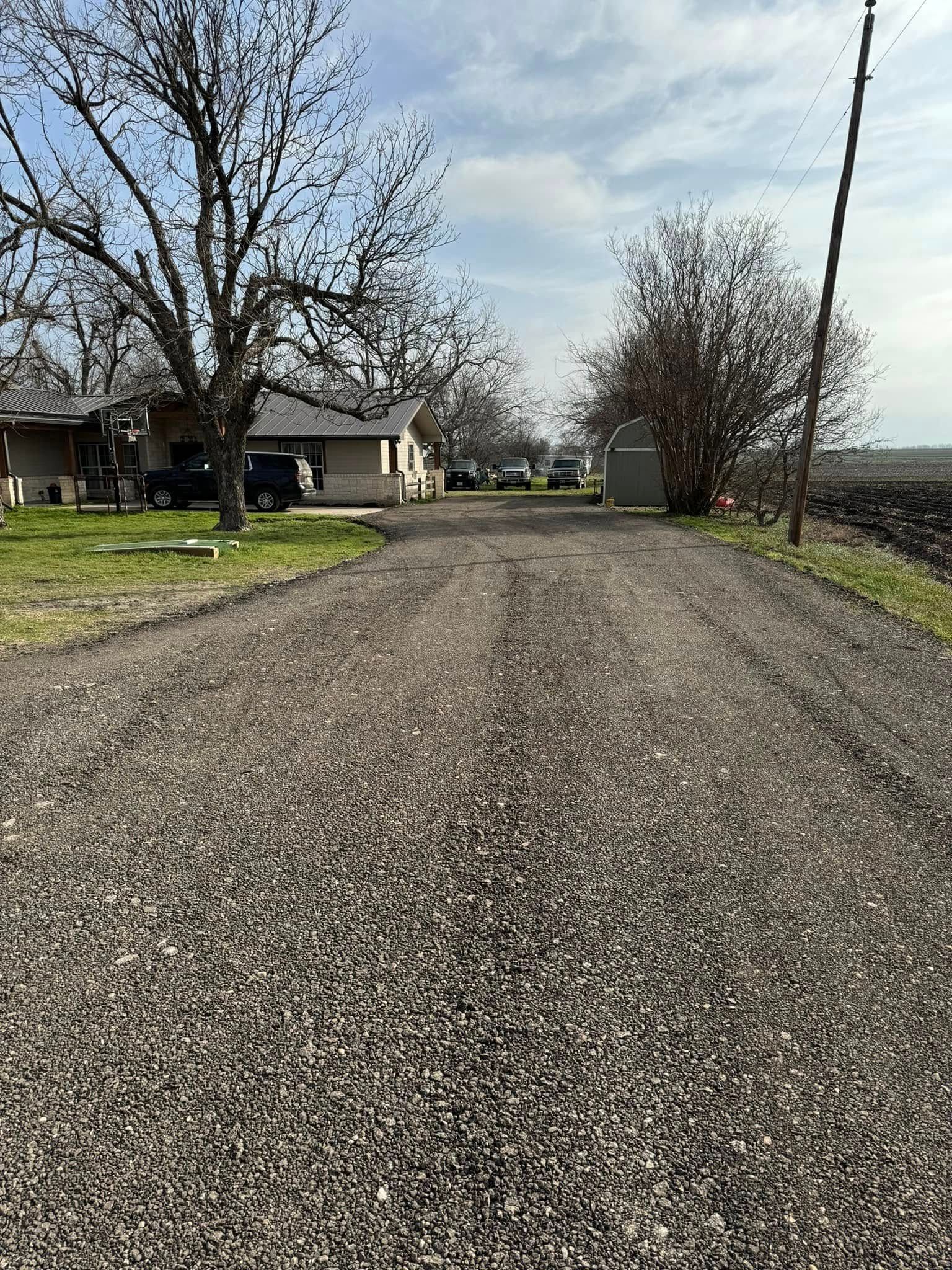 Gravel driveway leading to a small house under a cloudy sky, with trees and a shed on the sides.