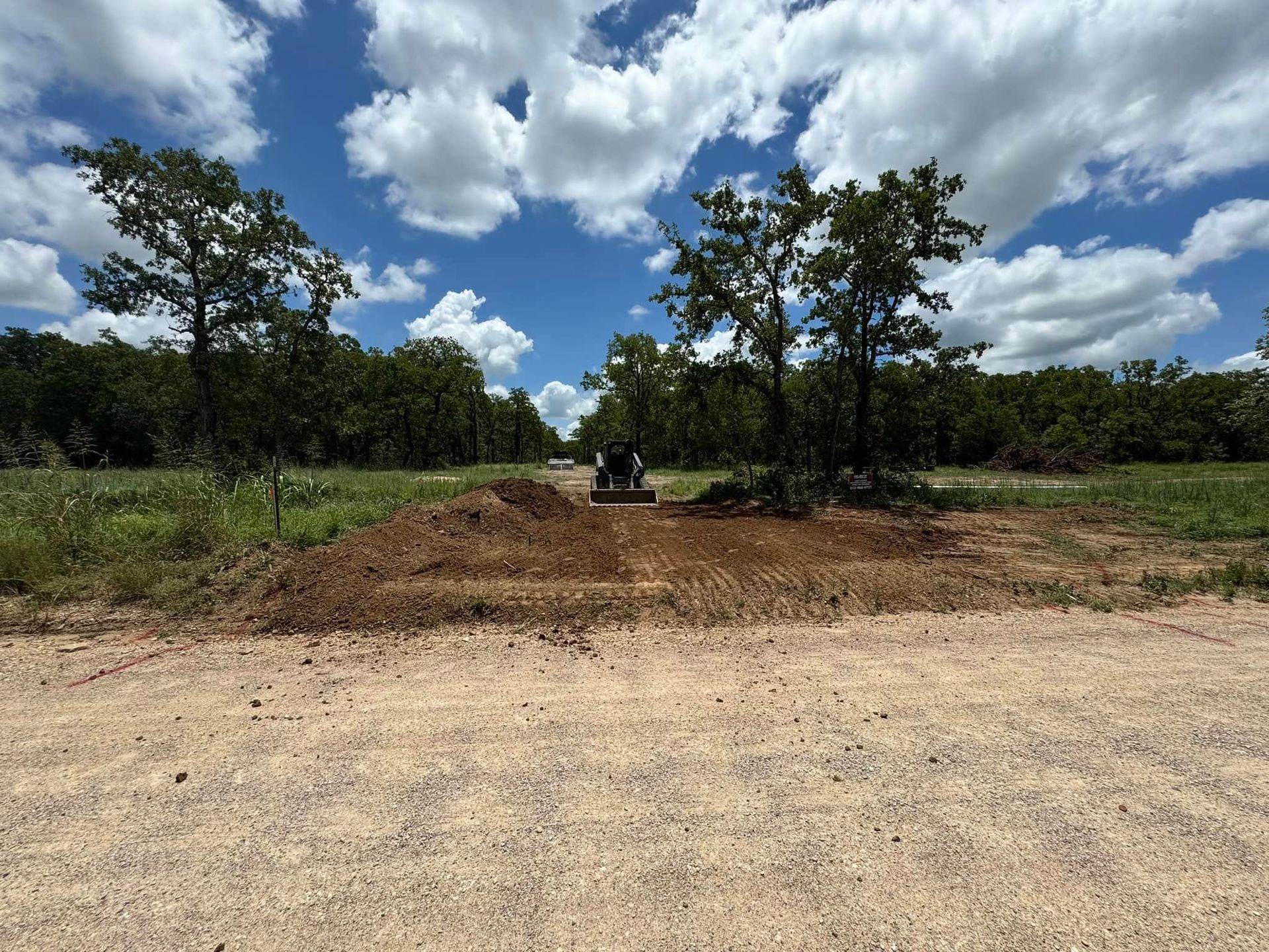 Dirt road with trees and a small pile of dirt under a bright blue sky with puffy clouds.
