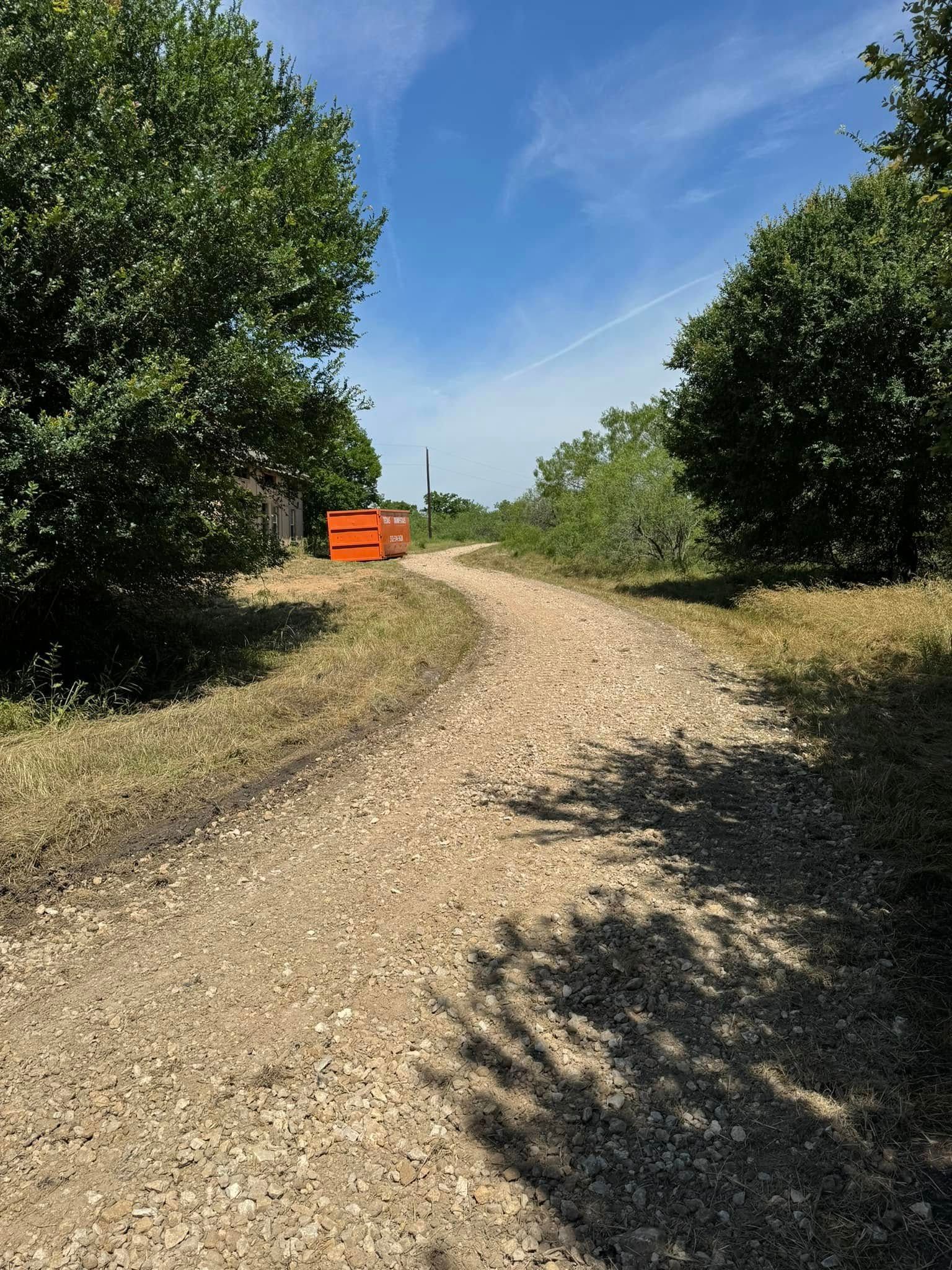 Gravel path lined by trees, leading towards orange container under a bright blue sky.