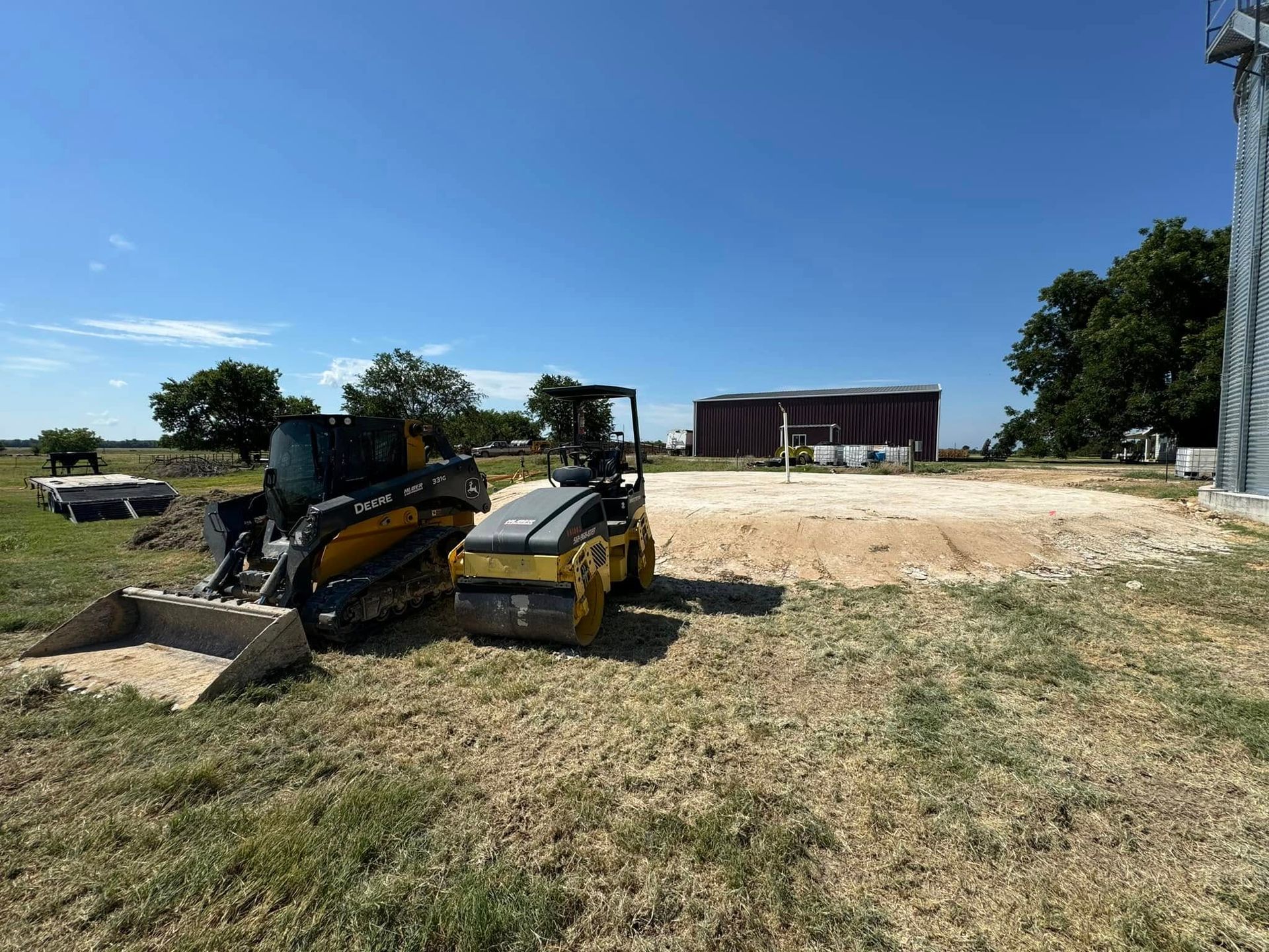 Construction site with a small bulldozer and compactor; blue sky, dirt, and a grain silo visible.