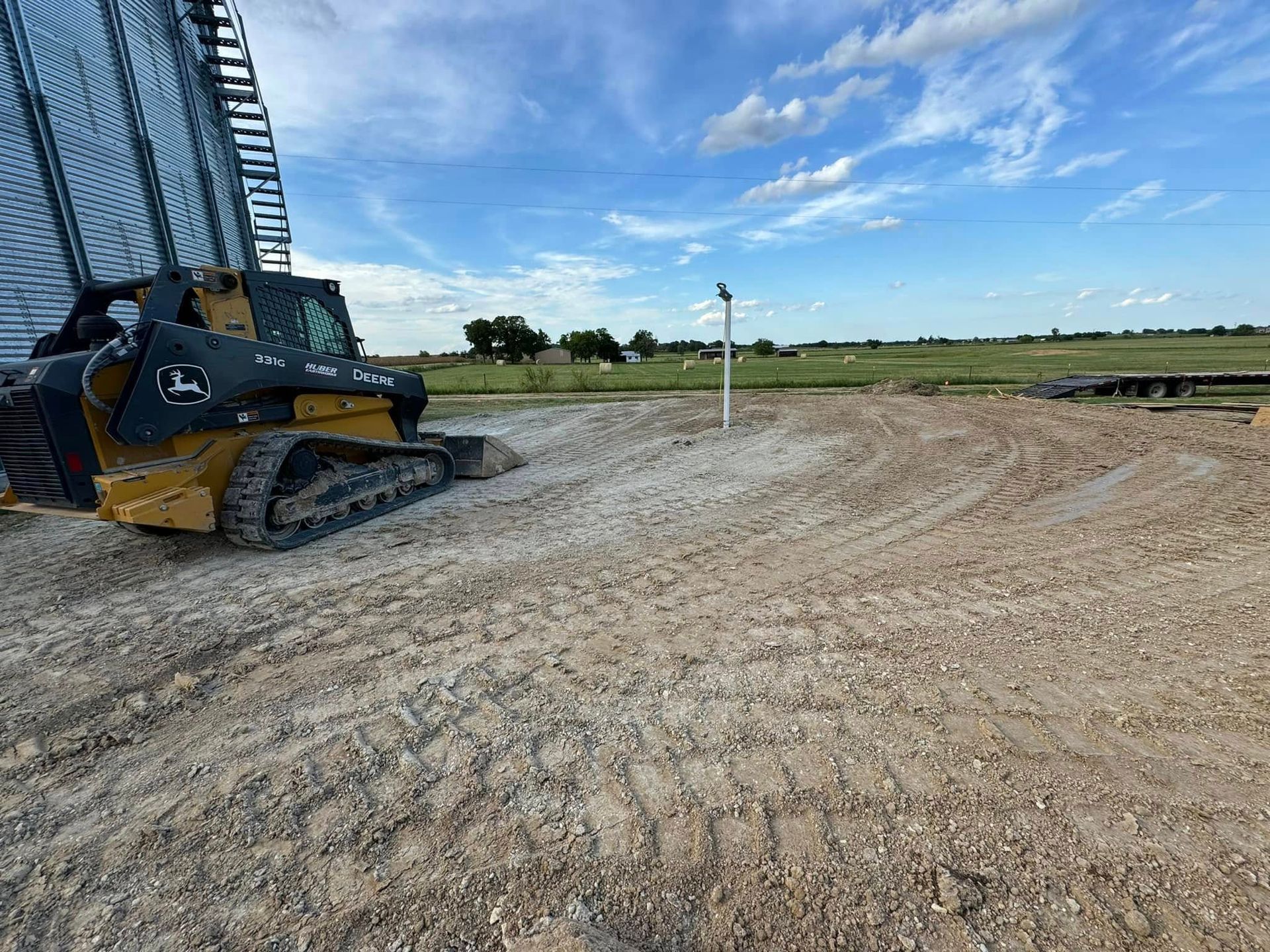 A John Deere skid steer on a dirt field near a grain silo under a blue sky.