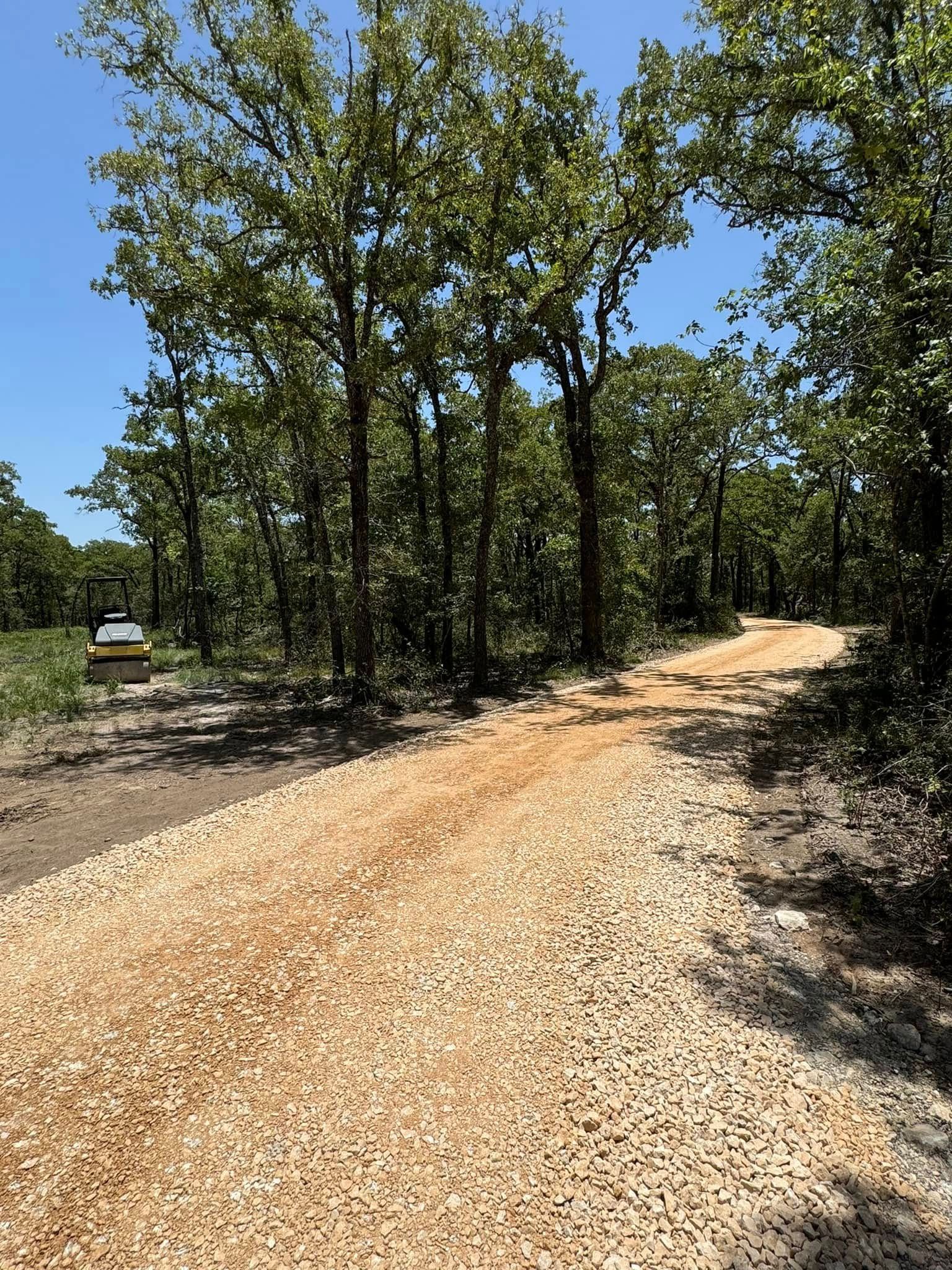 Gravel road leading into a wooded area with trees and a bright blue sky.
