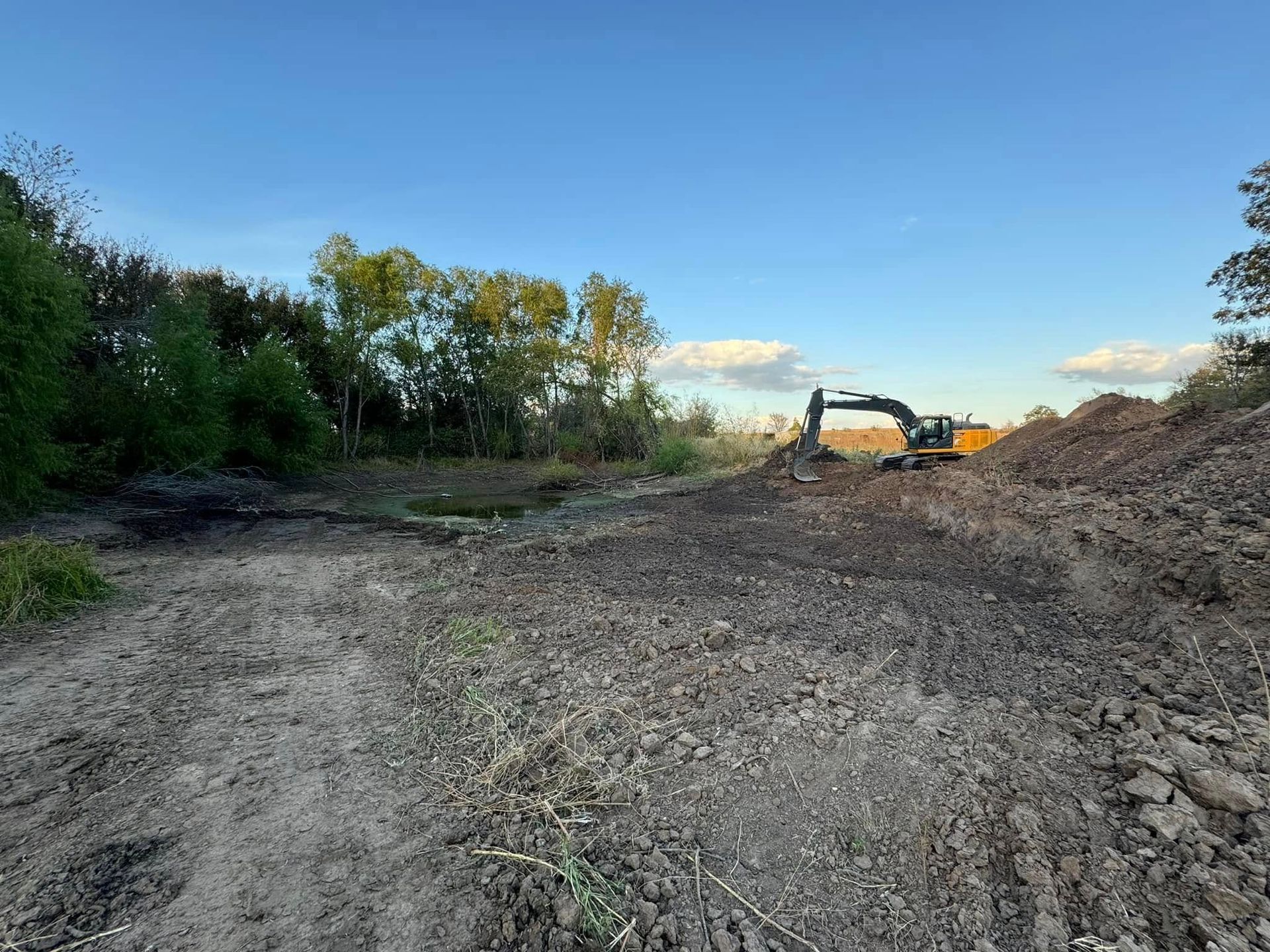 An excavator on a dirt pile near trees, with a clear blue sky above.