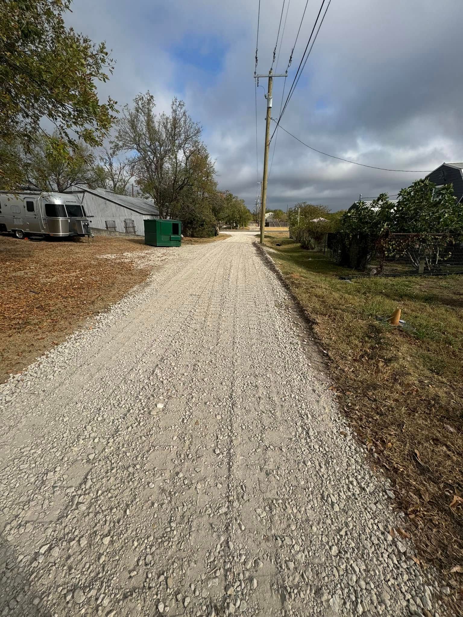 Gravel path through a rural area, next to trees and a utility pole. An Airstream trailer is parked nearby.