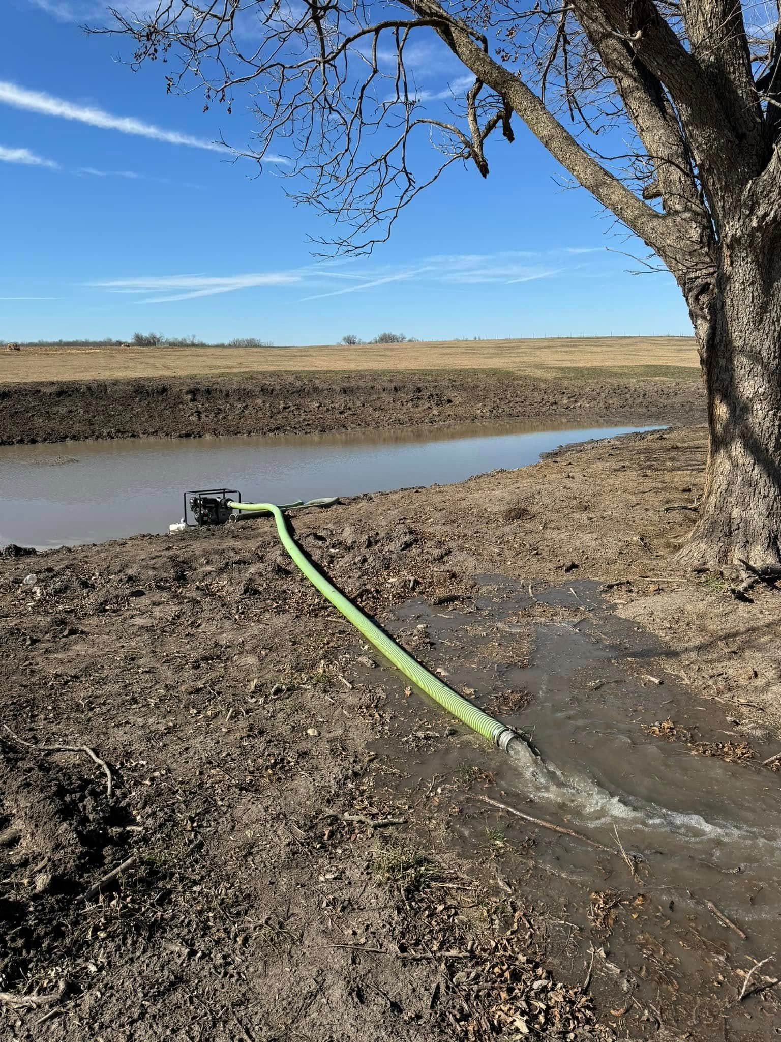 A pump with green hose discharges water into a muddy ditch. A tree is on the right, blue sky in the background.