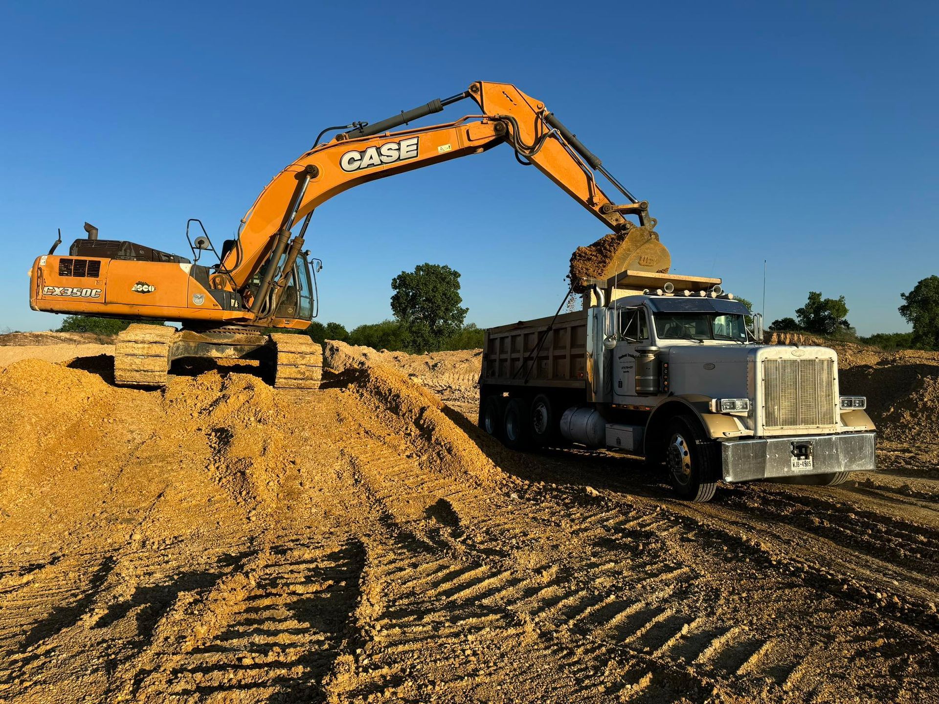 An orange Case excavator loading dirt into a white dump truck on a sunny day.