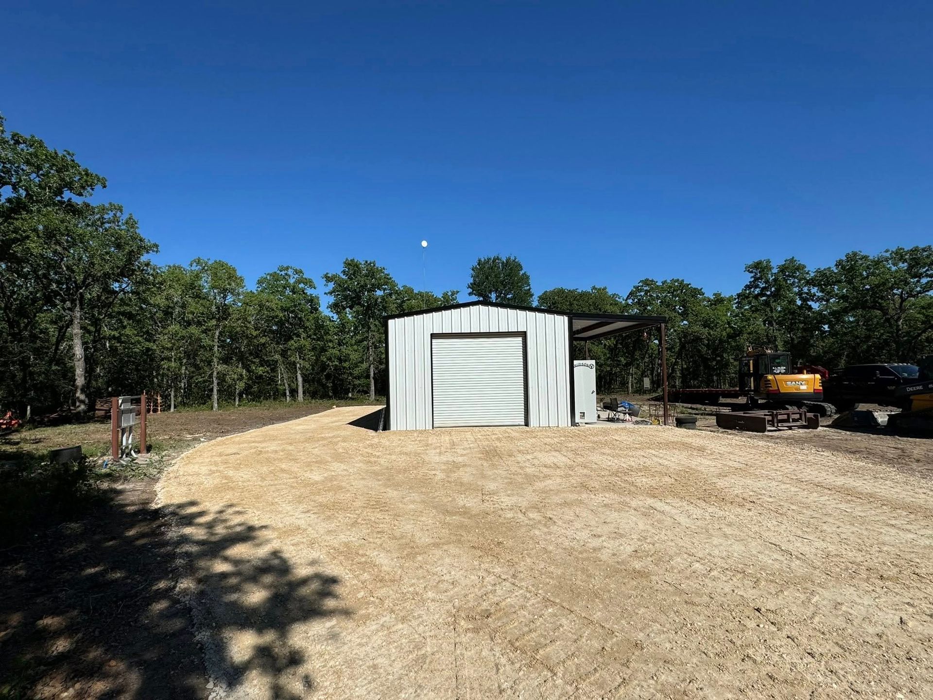 Gravel driveway leading to a white metal shed in a wooded area under a bright blue sky.
