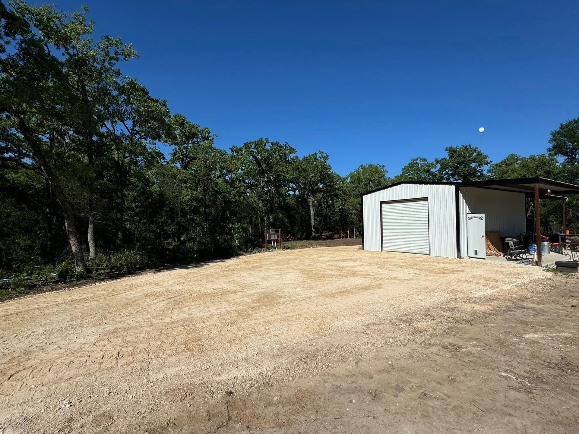 Gravel driveway with a white metal garage and trees under a clear blue sky.