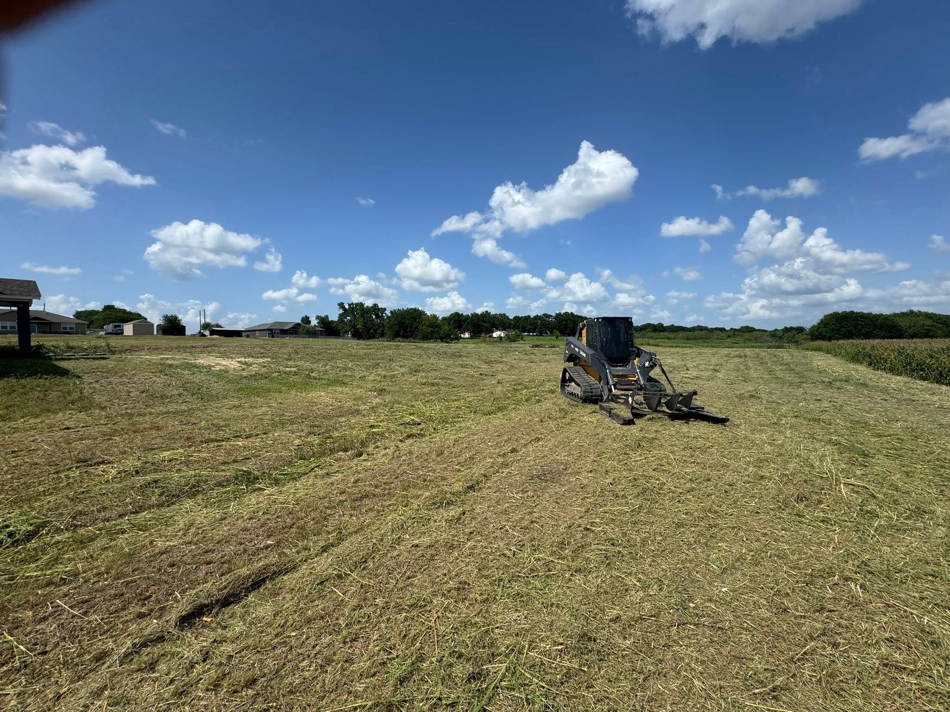 Skid steer mowing tall grass on a field under a partly cloudy blue sky.