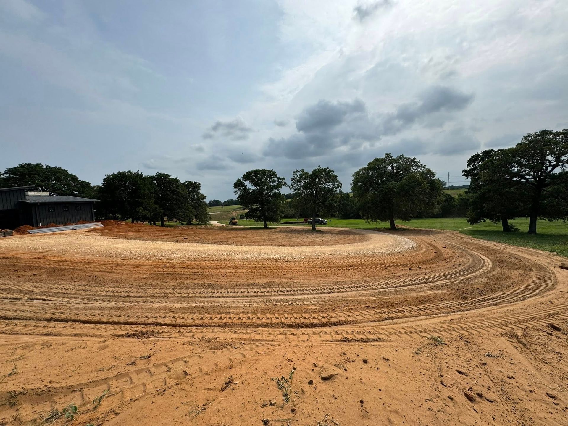 Dirt racetrack under cloudy sky, with trees and building visible in the background.