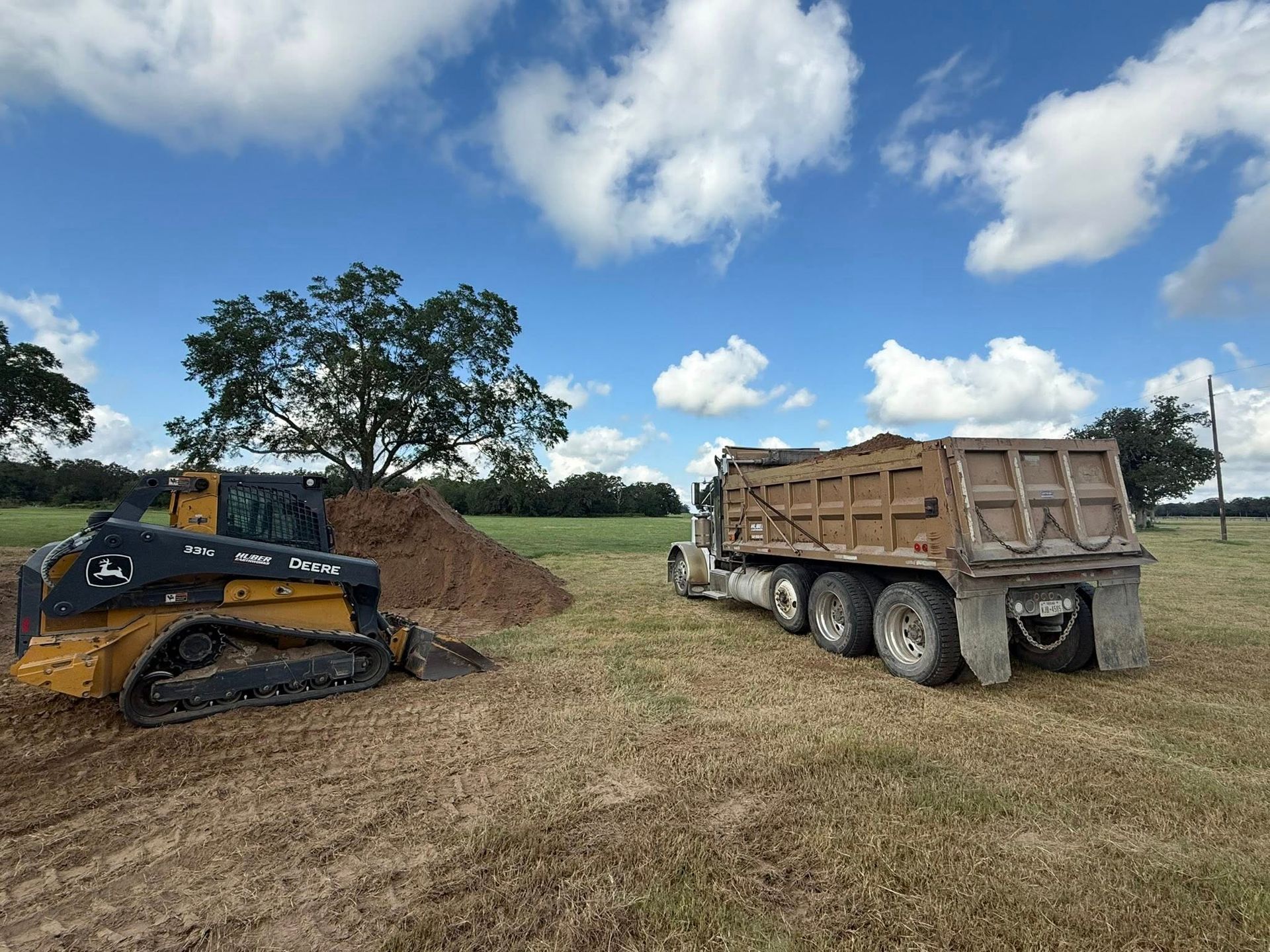 A John Deere track loader scoops dirt near a dump truck in a field under a cloudy sky.