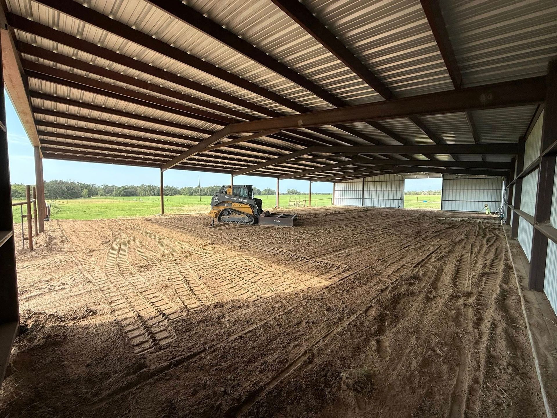 Inside a large open-air metal building, a compact tractor works on a dirt floor.