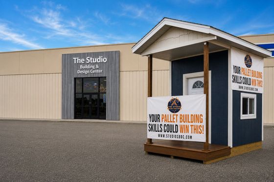 Small white kiosk with signs outside The Studio building on a gravel lot under a blue sky.