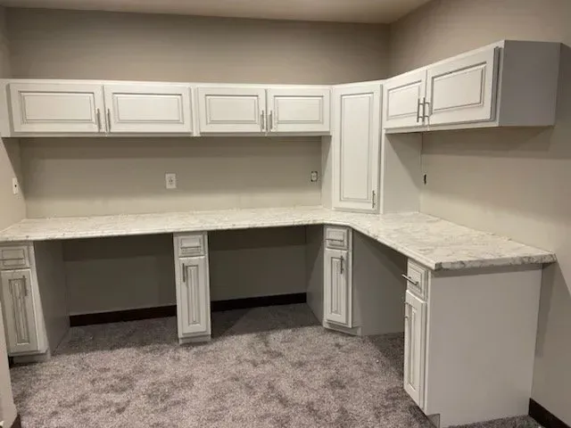 There is a large desk in the middle of the room with white cabinets.