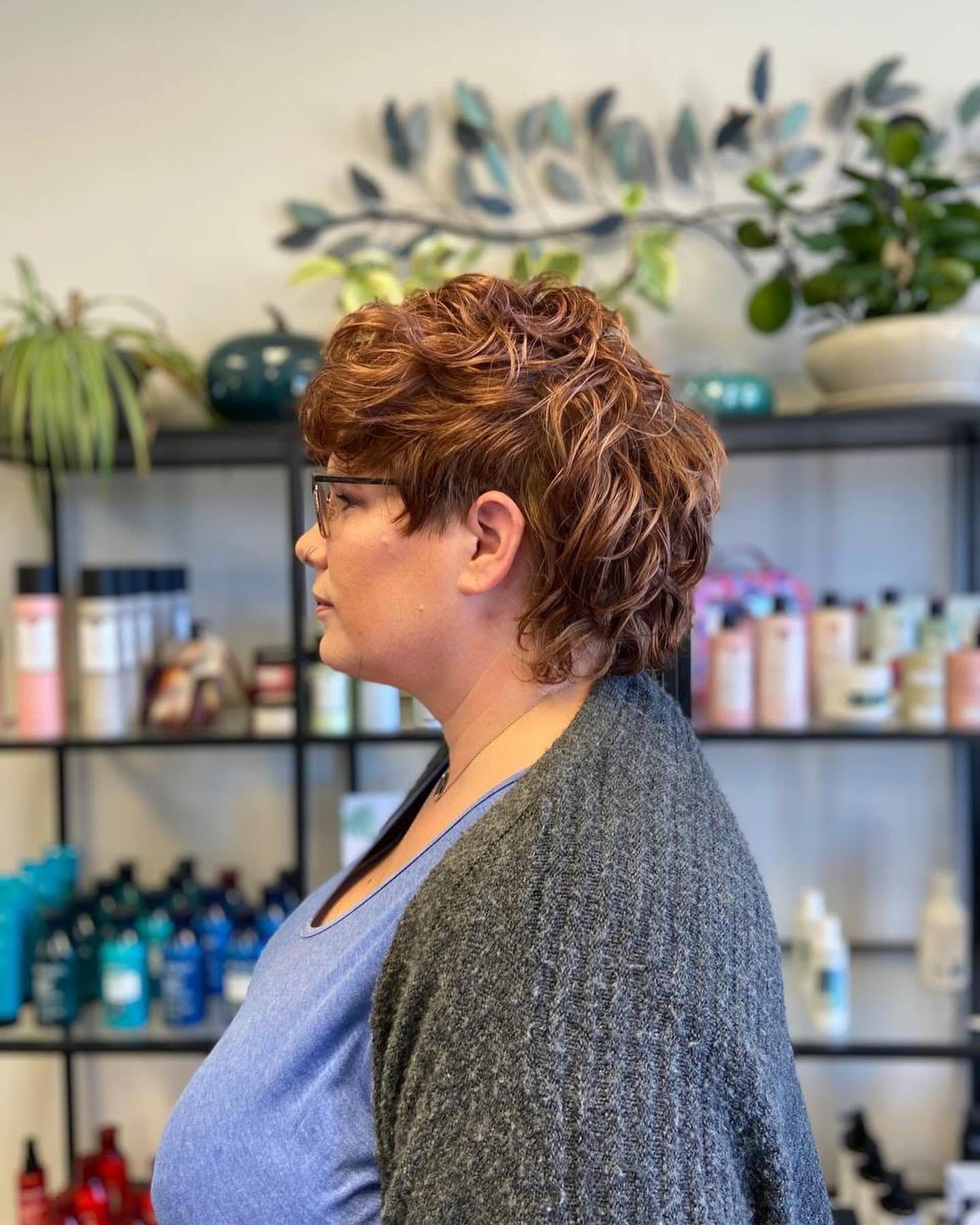 A woman with short red hair and glasses is standing in front of a shelf full of hair products.