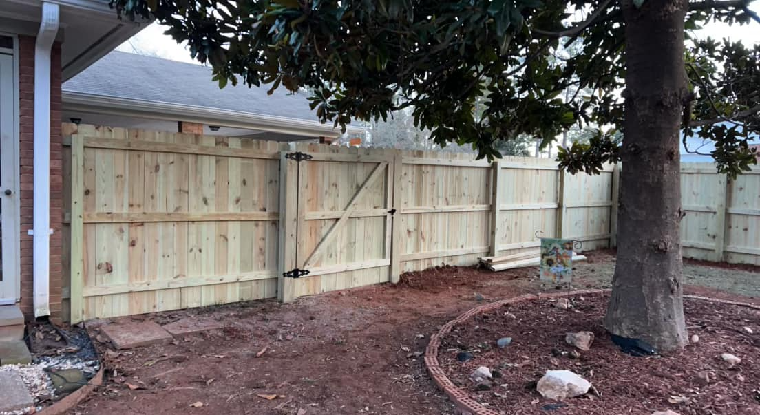 Wooden fence enclosing a yard, with gate and tree; dirt and mulch ground cover.