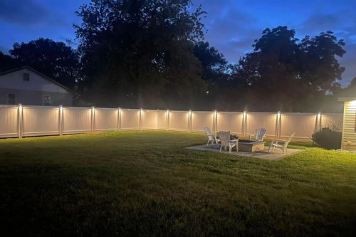 Backyard at dusk, lit fence with lights along the top, fire pit, chairs on the patio, and green grass.