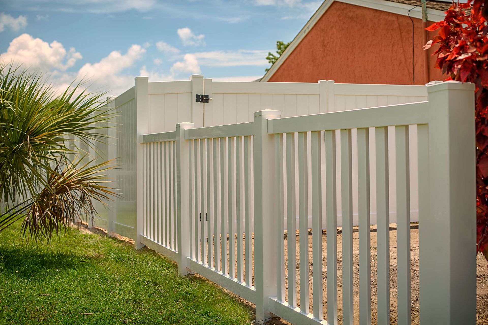 White closed top picket fence with vertical slats bordering a yard, with green grass and blue sky.