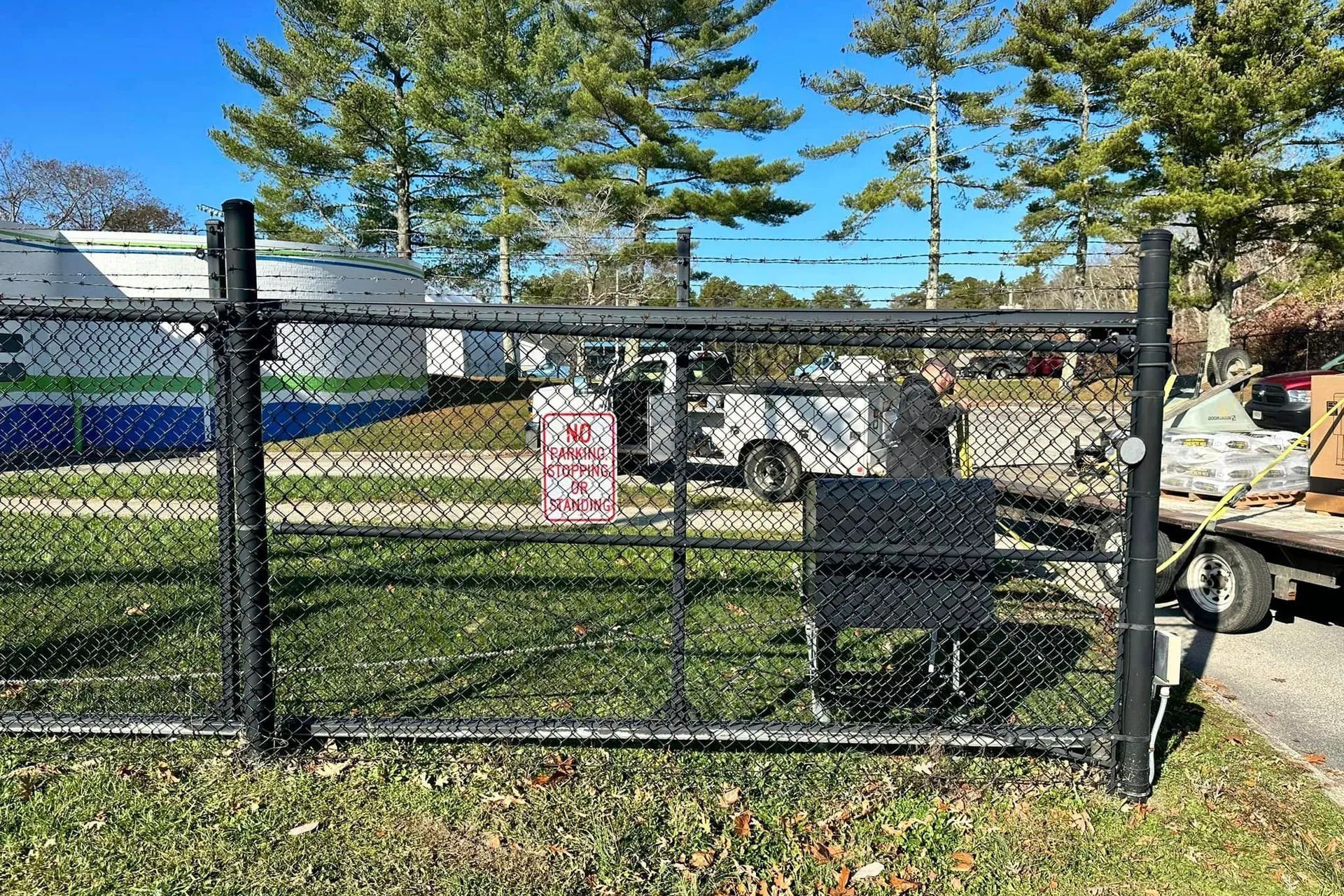 Black chain-link fence encloses a grassy area. A truck with open doors is behind the fence with a sign. Trees in the background.