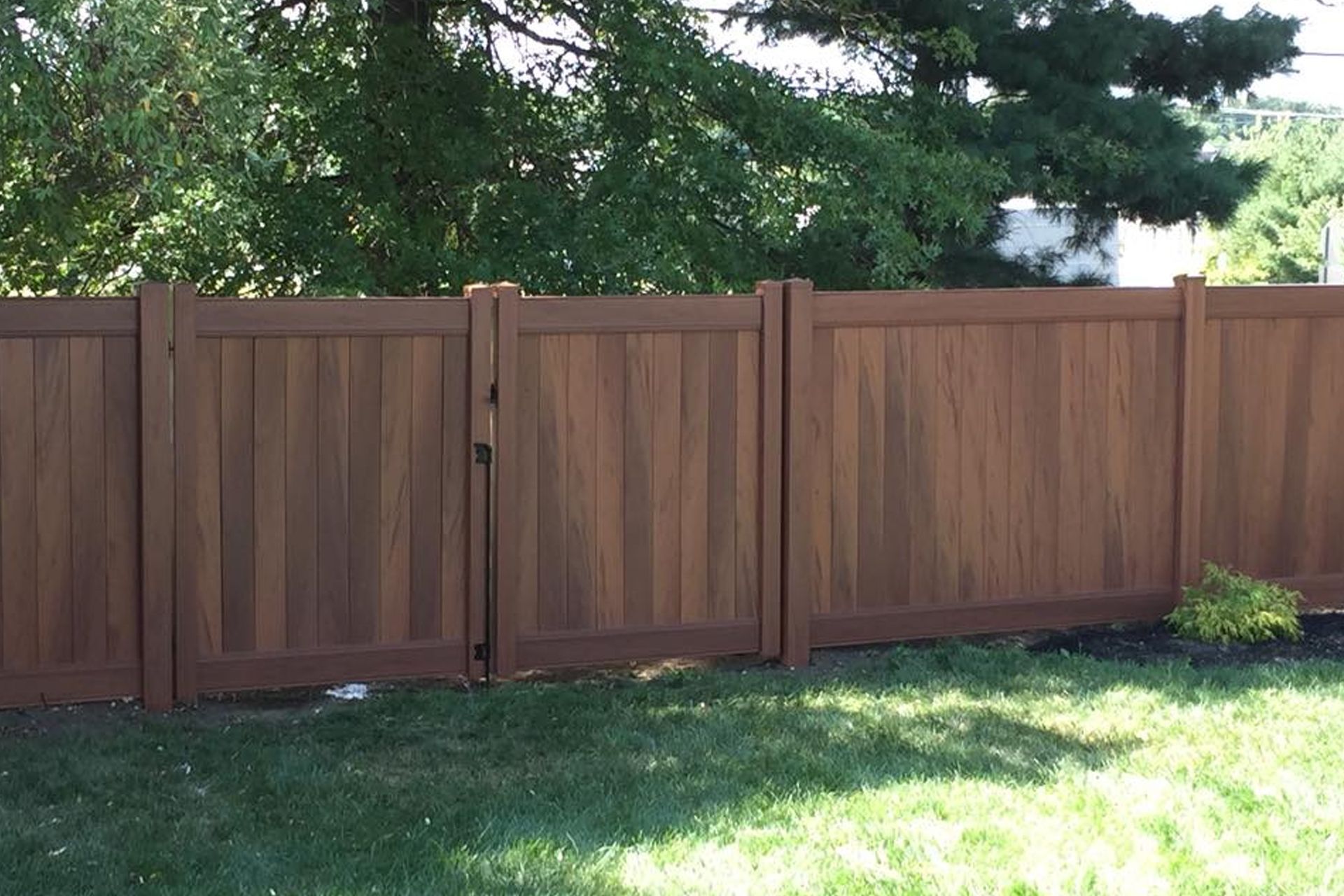 Brown wood-look fence with gate in grassy yard, trees in background.
