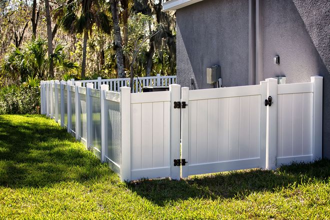 Black chain-link fence encloses a grassy area. A truck with open doors is behind the fence with a sign. Trees in the background.