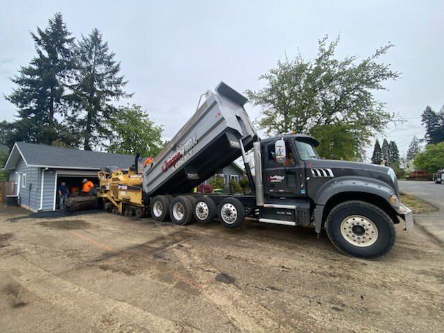 A dump truck is driving down a dirt road next to a house.