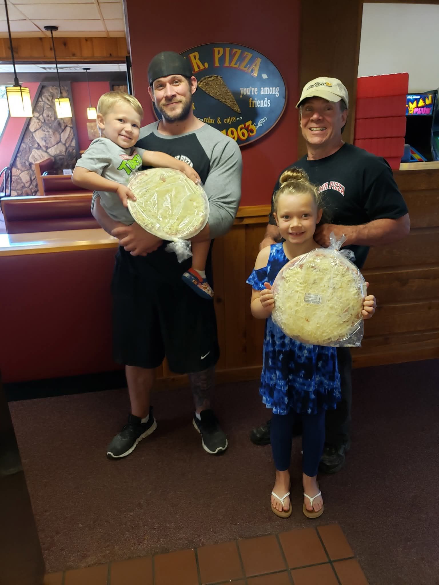 Two adults and two children holding large pizzas inside a restaurant, smiling at the camera.