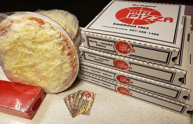 Stacked pizza boxes beside a large wrapped pizza and red packaged items on a countertop