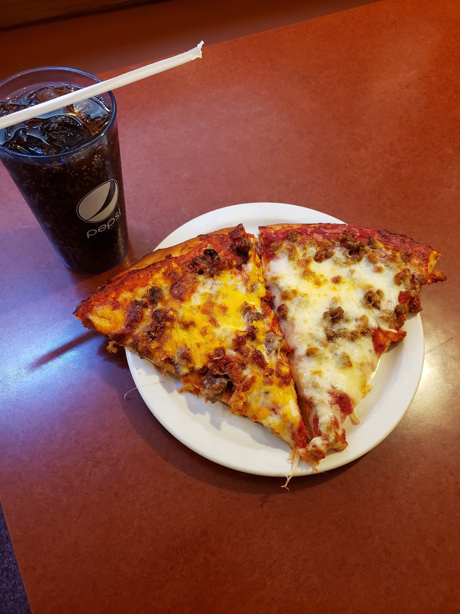 Two slices of pizza on a white plate beside a dark soda with a straw on a brown table