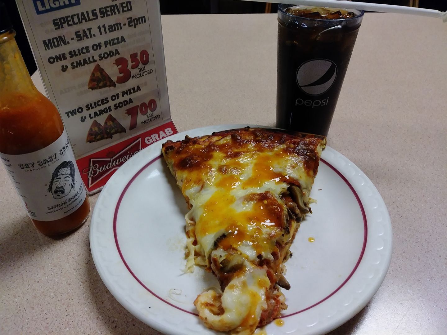 Slice of cheesy pizza on a plate with a soda, hot sauce bottle, and menu in a restaurant setting