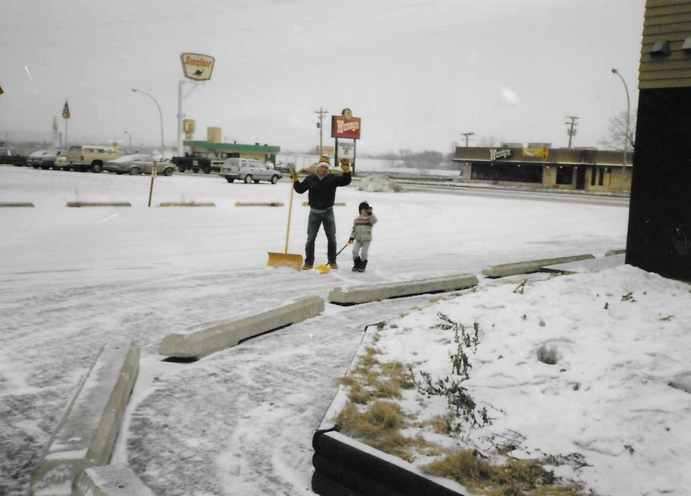 Person and child standing in a snowy parking lot, with a shovel and winter-covered ground.