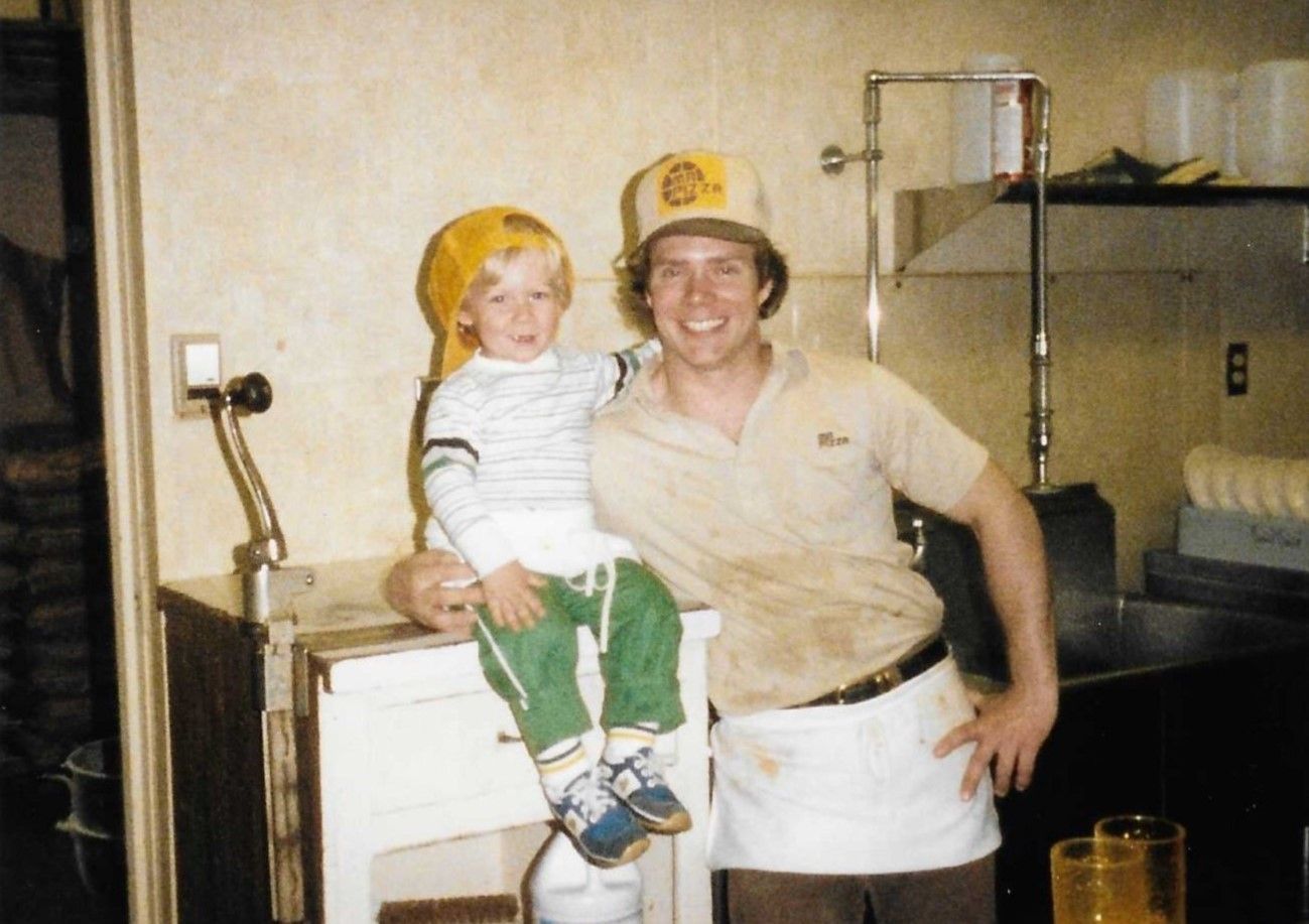 Man in a white apron holding a child in a kitchen, both wearing yellow hairnets, smiling.
