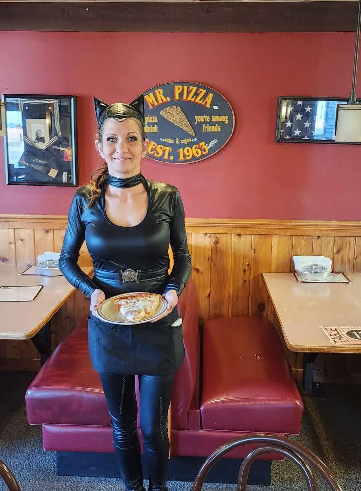 Woman in black outfit holding a plate, standing in a red booth inside a pizza restaurant.