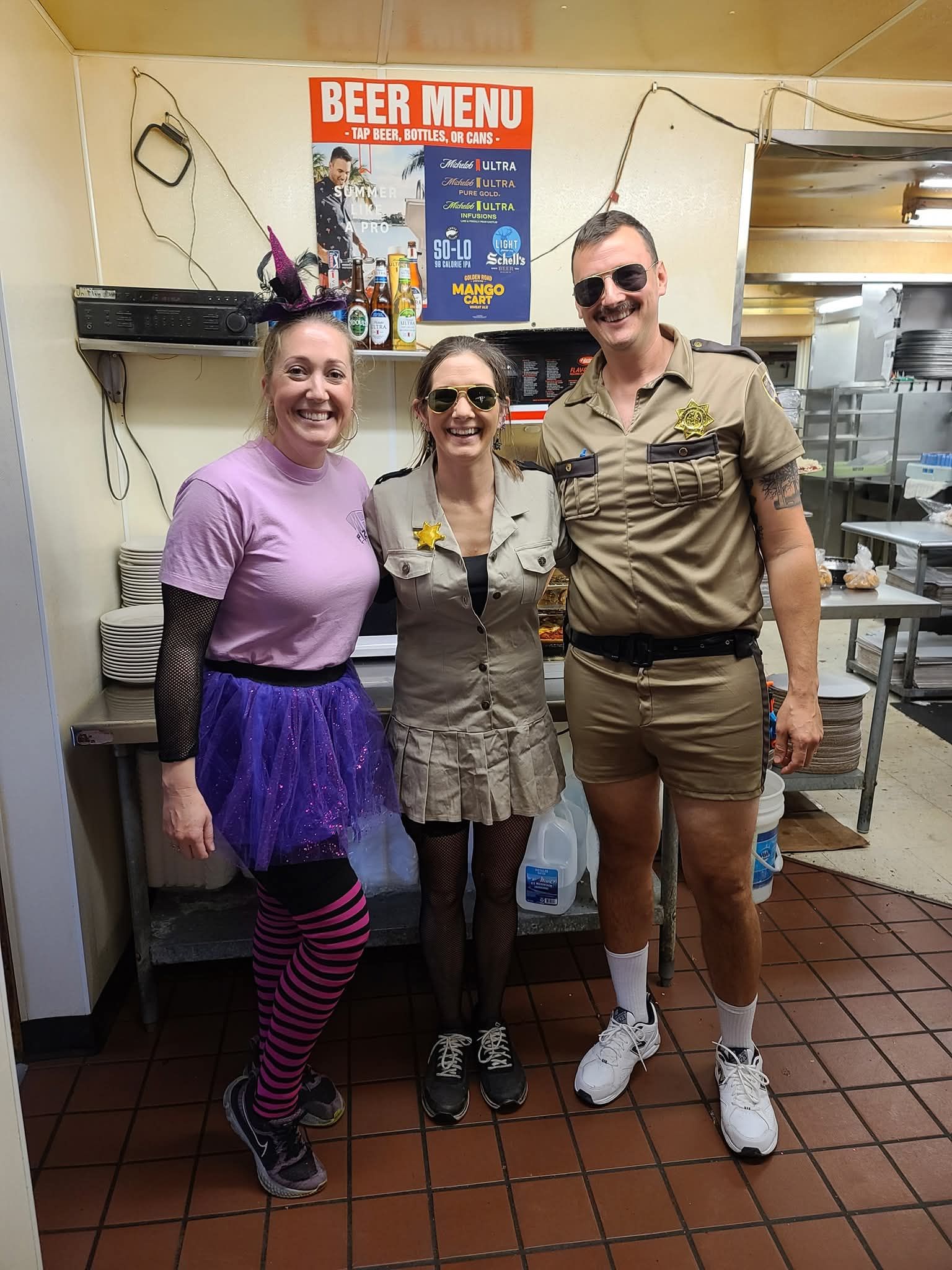 Three people smiling in costume, standing indoors under a beer menu sign.