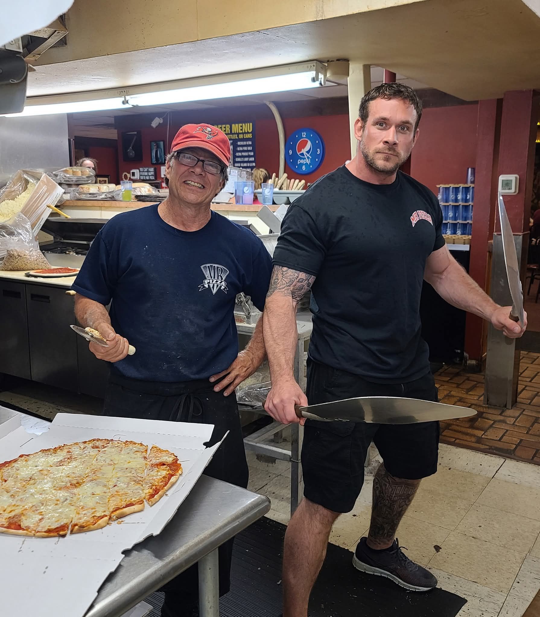 Two men in a pizza shop hold large pizzas, with one pizza on a prep table in the foreground.