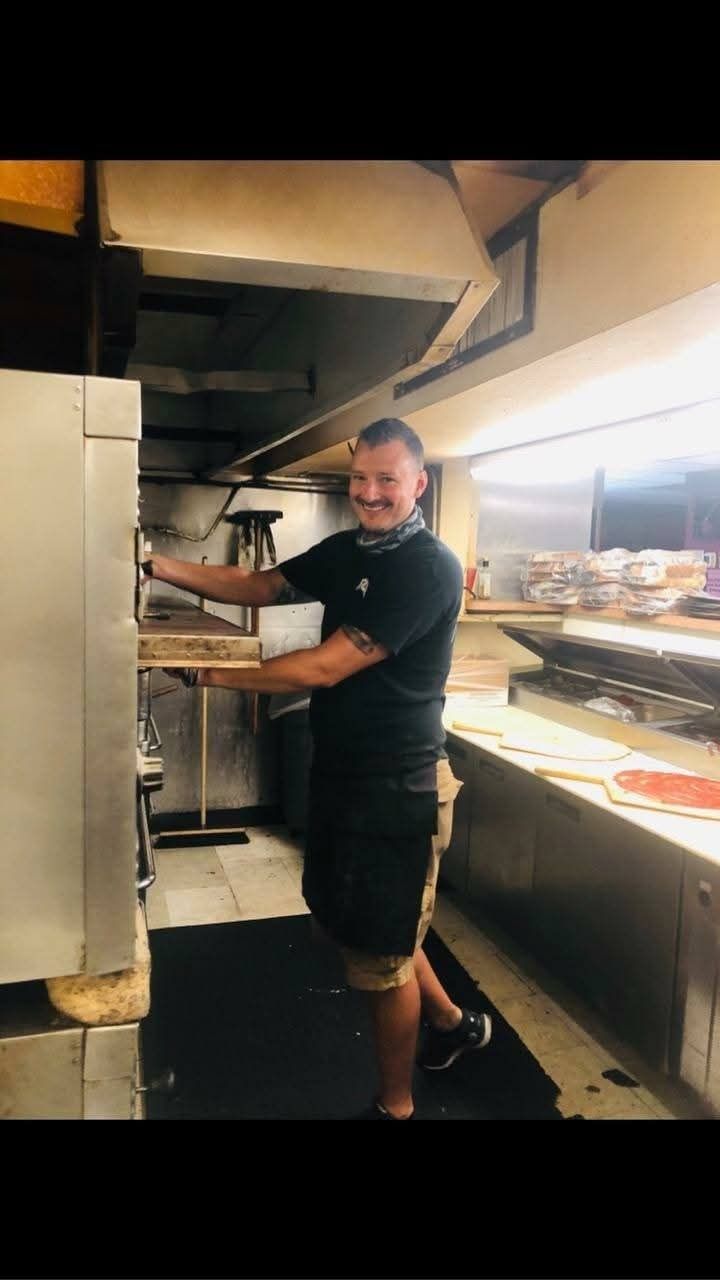 Chef in black uniform smiling in a commercial kitchen, holding equipment beside a pizza prep counter.