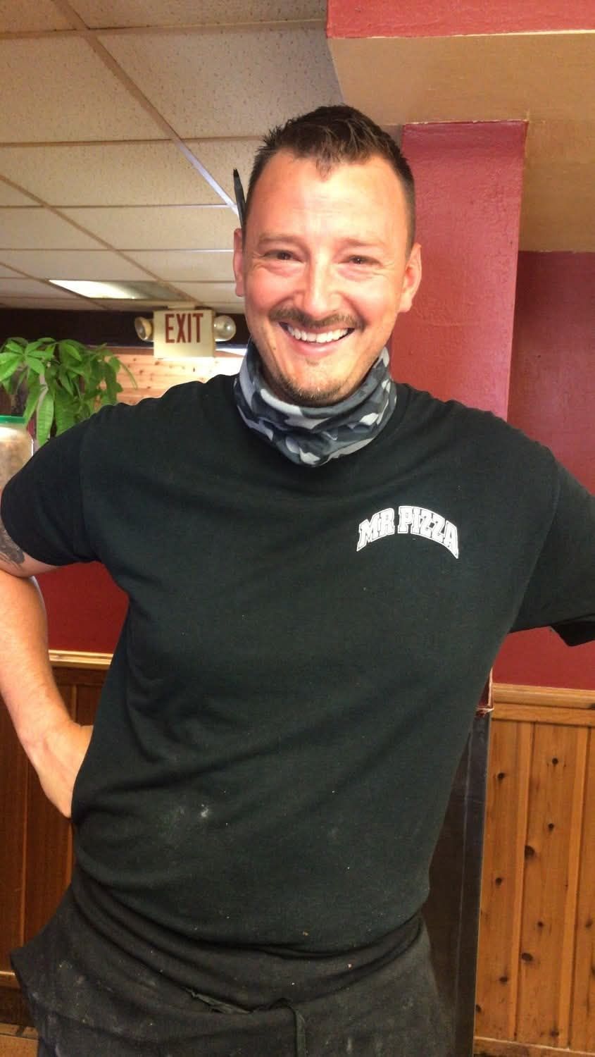 Smiling man in a black T-shirt and neck gaiter stands indoors with hands on hips.