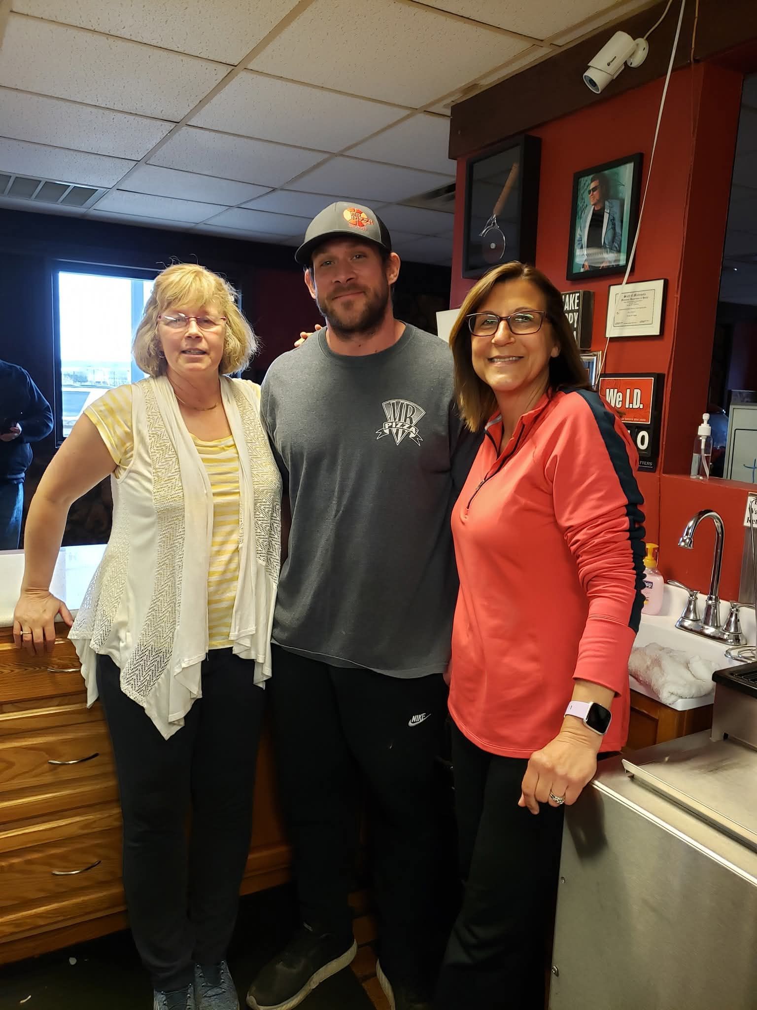Three people posing indoors, smiling in a restaurant or bar with red walls and a countertop.