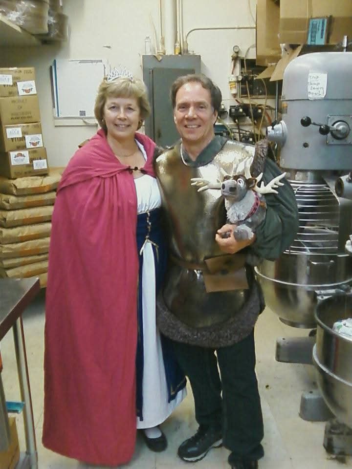 Two people in medieval costumes stand in a workshop, one holding a small dog.