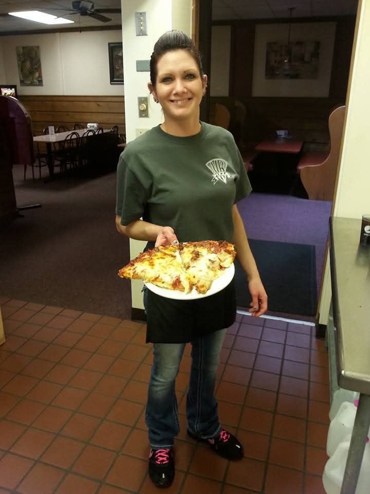 Person holding a slice of pizza, smiling in a dimly lit dining area.