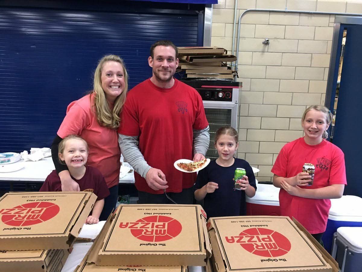 Group of five people in red shirts standing behind pizza boxes in a warehouse, holding plates and drinks.