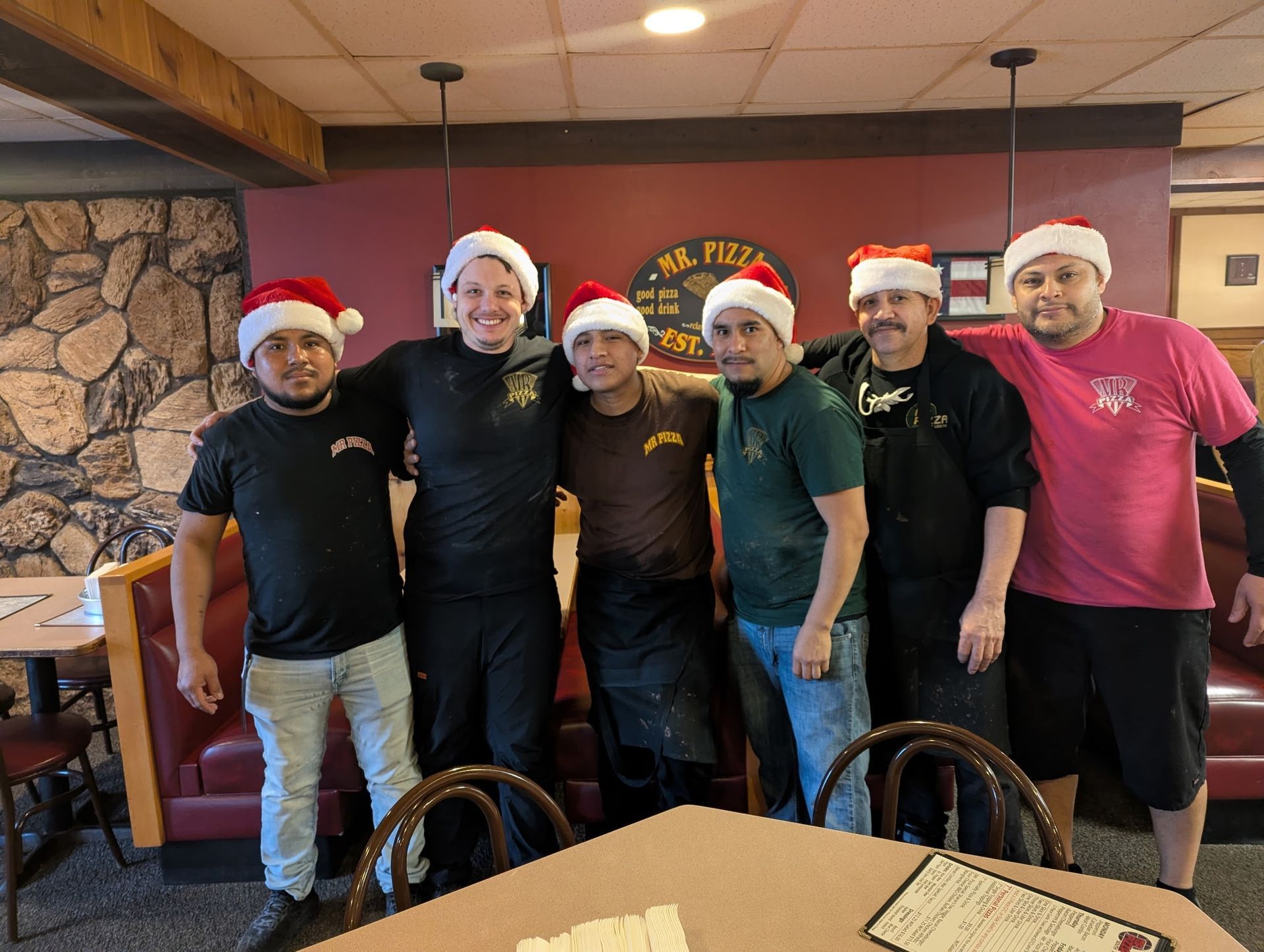 Six people in Santa hats posing together in a restaurant booth area, smiling at the camera.